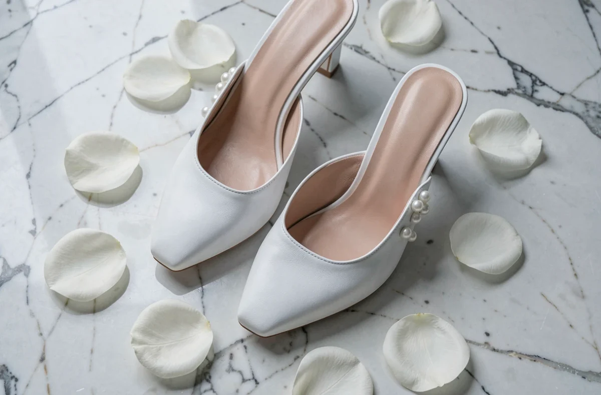 Gros plan sur des mules blanches à petit talon en cuir avec ornements de perles sur marbre et pétales de roses